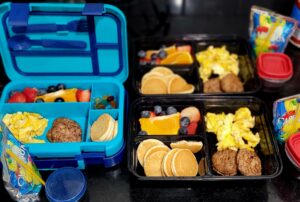 School morning breakfast boxes with scrambled eggs, sausage patties, mini pancakes, fruit, and juice pouches in divided containers